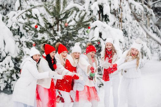 A large group of girls with glasses of champagne in their hands stands in the winter forest.Girls in red and white clothes with new year's drinks in a snow-covered forest