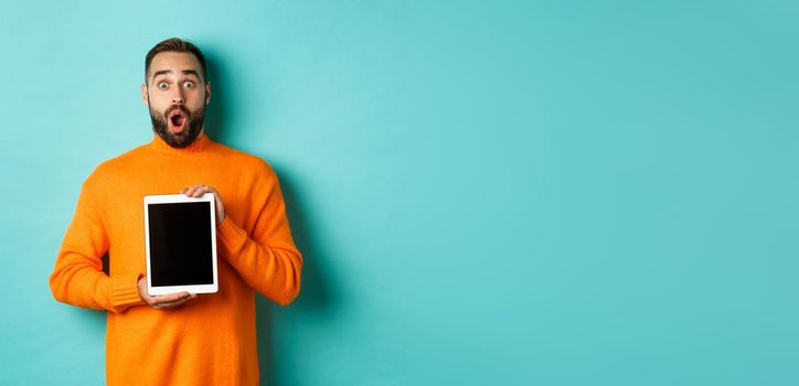 Technology. Surprised bearded man drop jaw, staring at camera and showing digital tablet screen, standing againt light blue background.