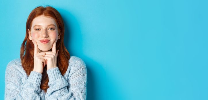 Close-up of cute and silly girl with red hair poking cheeks, showing dimples and smiling, standing against blue background.