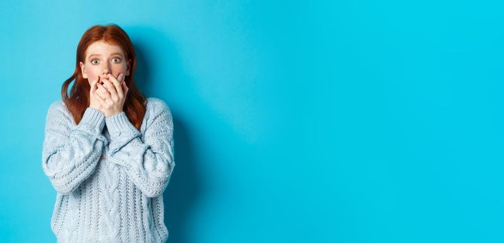 Shocked redhead girl in sweater, staring at camera scared and gasping, covering mouth with hands, standing against blue background.