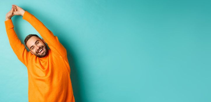 Image of handsome young man stretching hands and smiling after good rest, standing in orange sweater over light blue background.