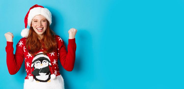 Happy holidays and Christmas concept. Cheerful redhead girl in santa hat and xmas sweater, winning and celebrating victory, raising hands up satisfied, triumphing.