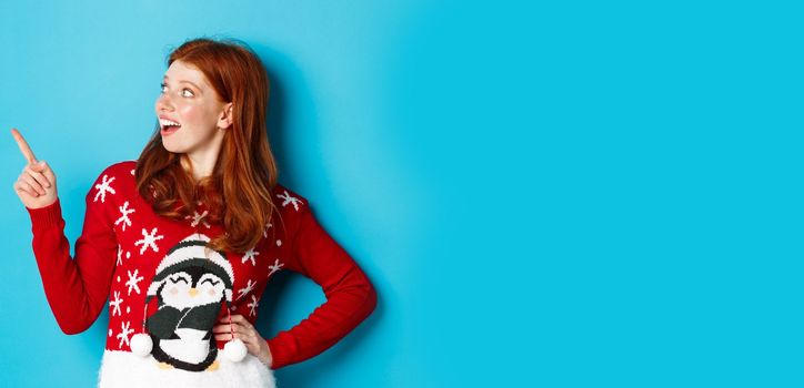 Winter holidays and Christmas Eve concept. Happy and cute redhead girl in xmas sweater, looking and pointing at upper left corner amazed, standing over blue background.