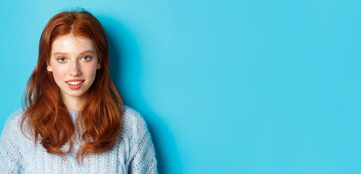 Close-up of young cute redhead girl smiling at camera, standing against blue background.