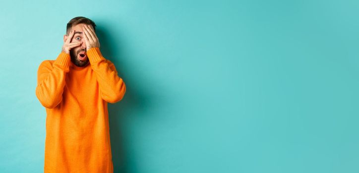 Curious man peeking at something, shut eyes but looking through fingers with amazement and interest, standing over light blue background.