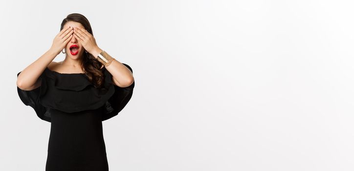 Fashion and beauty. Beautiful woman in black dress and red lipstick, covering eyes and smiling, waiting for surprise, standing over white background.