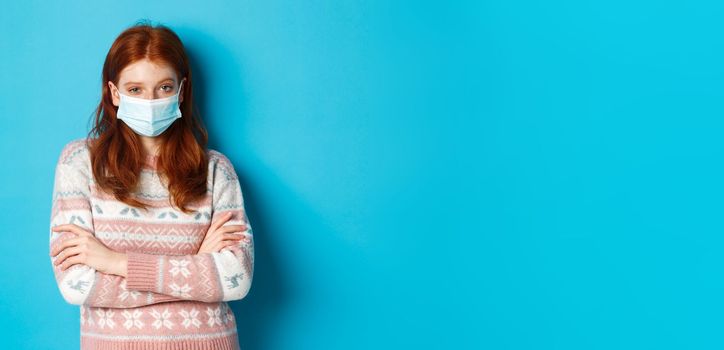 Winter, covid-19 and quarantine concept. Skeptical redhead girl in sweater and medical mask, cross arms on chest and staring with disbelief, standing over blue background.