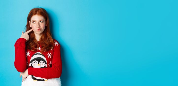 Winter holidays and Christmas Eve concept. Pretty redhead girl in xmas sweater, touching cheek thoughtful and smiling, making choice, looking at upper left corner and thinking, blue background.