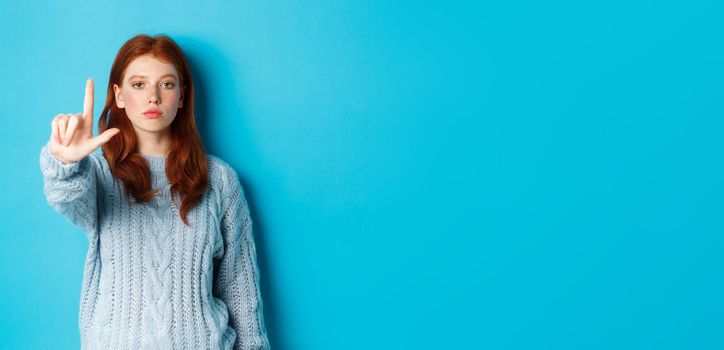 Serious redhead girl in sweater showing taboo gesture, extending one finger, shaking forefinger to disapprove, disagree and forbid something, blue background.