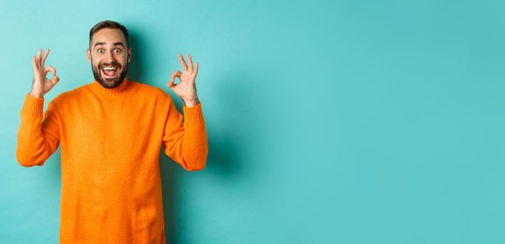 Impressed man praising something awesome, showing okay signs and looking amazed, standing in orange sweater at light blue background.