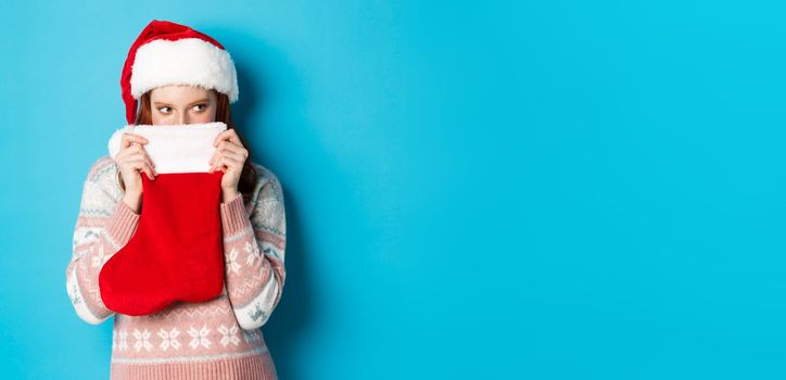 Cute girl cover face with christmas stocking, staring right with cunning gaze, standing in Santa hat and celebrating winter holidays, blue background.