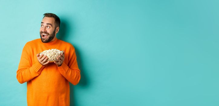 Excited and happy man watching tv and holding bowl of popcorn, looking left and smiling pleased, standing over blue background.