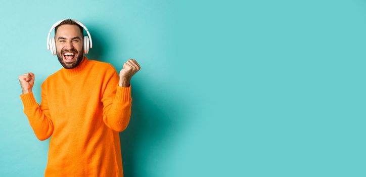 Happy adult man in orange sweater, looking up and listening music in headphones, standing over blue background.