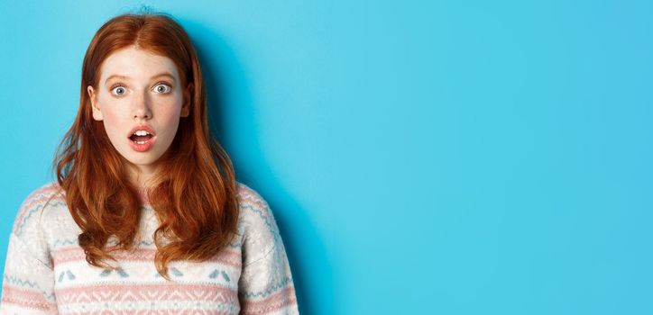Close-up of shocked redhead girl drop jaw in awe, staring with amazement at camera, standing against blue background.