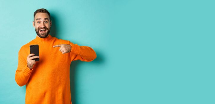 Excited man pointing at mobile phone, showing internet promo, standing over turquoise background, concept of online shopping.