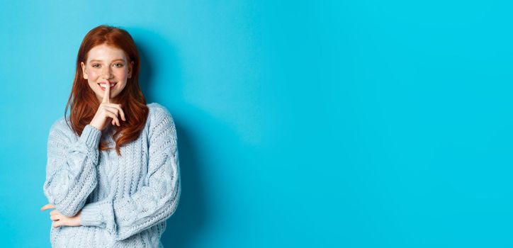 Pretty redhead teenager hushing and smiling, telling a secret, standing in sweater against blue background.