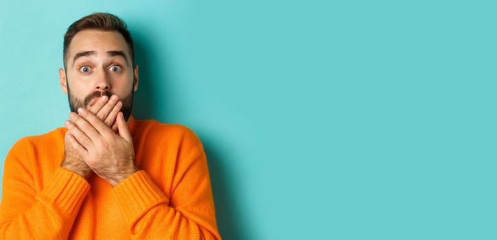 Close-up of shocked caucasian man covering his lips and mouth, staring astounded at camera, standing against turquoise background.