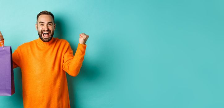 Happy man holding purple shopping bag and rejoicing, got discount and celebrating, standing over turquoise background.