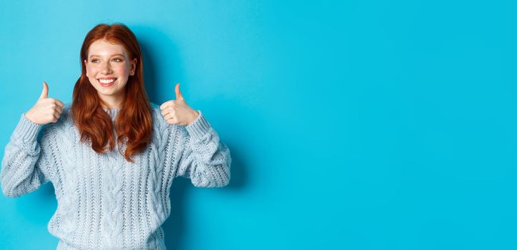 Happy smiling woman with red hair, showing thumbs up and looking left satisfied, praising good choice, agree and approve, standing over blue background.
