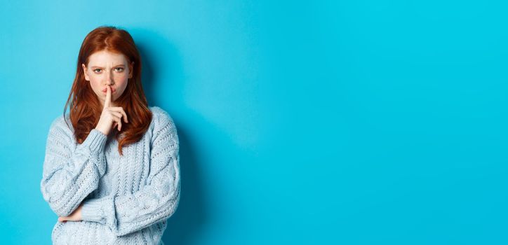 Angry redhead girl hushing at you, show taboo gesture, forbid to speak, standing over blue background in sweater.