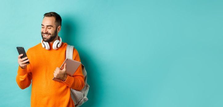 Handsome man student with headphones and backpack, holding digital tablet, reading message on mobile phone, standing against light blue background.