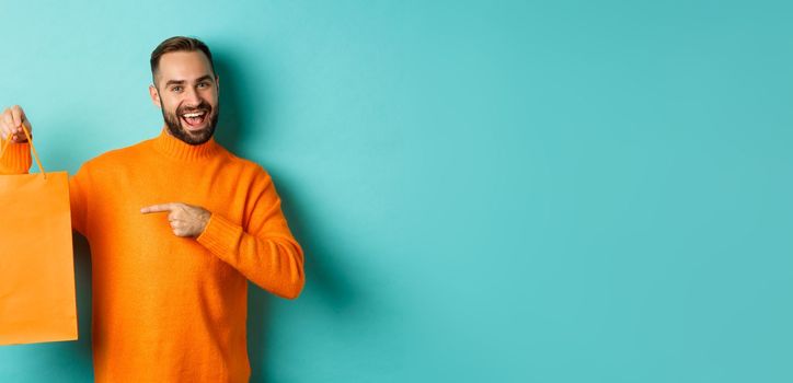 Satisfied male customer pointing at orange shopping bag, recommending store, smiling pleased, standing over turquoise background.