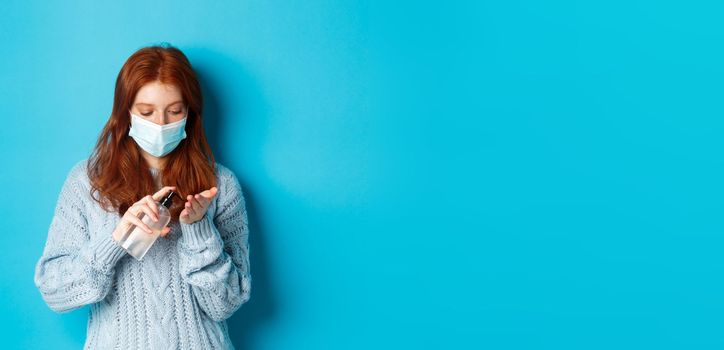 Winter, covid-19 and social distancing concept. Redhead female student in face mask clean hands with sanitizer, using antiseptic, standing against blue background.