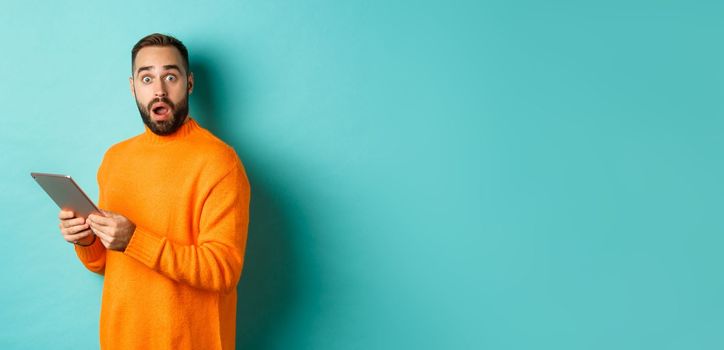 Image of male model in orange sweater using digital tablet, looking surprised, standing over light blue background.