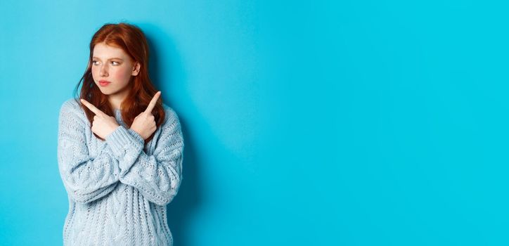 Indecisive redhead teenage girl making decision, pointing fingers sideways and looking left doubtful, standing in sweater against blue background.