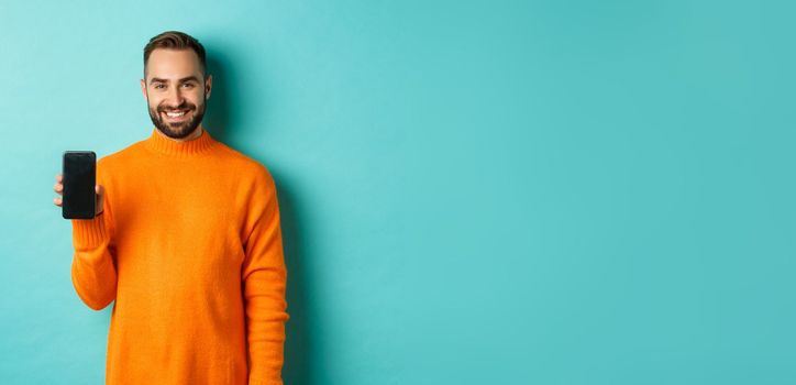 Handsome bearded guy in orange sweater, showing smartphone screen and smiling, showing promo online, turquoise background.