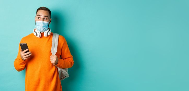 Young man in face mask using mobile phone, holding backpack, staring left amazed, standing against light blue background.