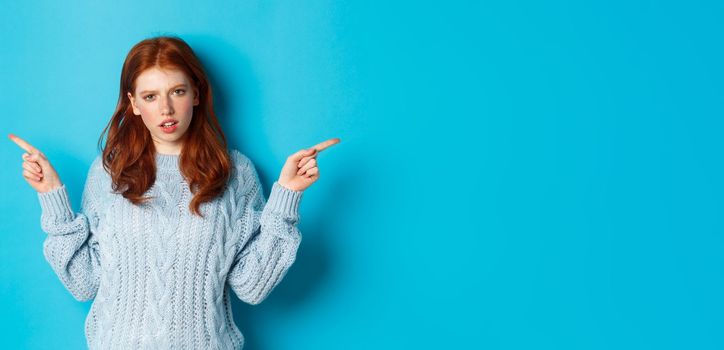 Confused redhead girl in sweater pointing fingers sideways, staring at camera doubtful, standing against blue background. Copy space