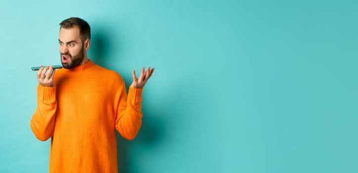 Angry man arguing on speakerphone, record voice message with mad face, standing over light blue background in orange sweater.