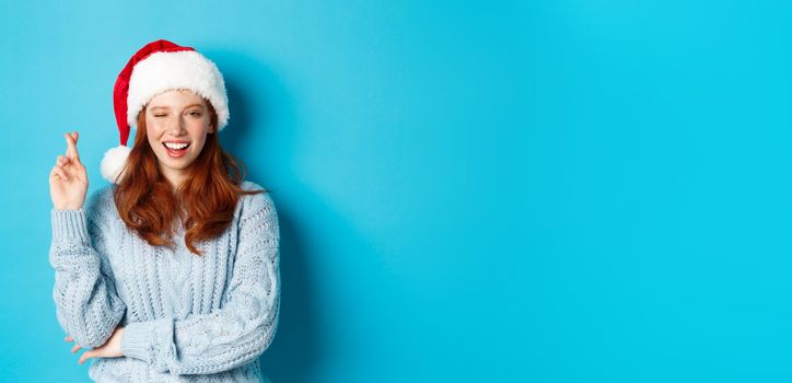 Winter holidays and Christmas Eve concept. Hopeful redhead girl in Santa hat, making wish on xmas, cross fingers for good luck and winking, standing over blue background.