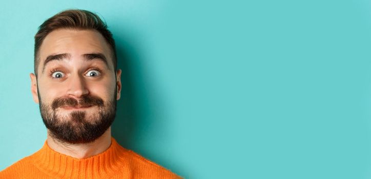 Headshot of funny bearded man showing faces and pouting, holding breath, standing in orange sweater against turquoise background.