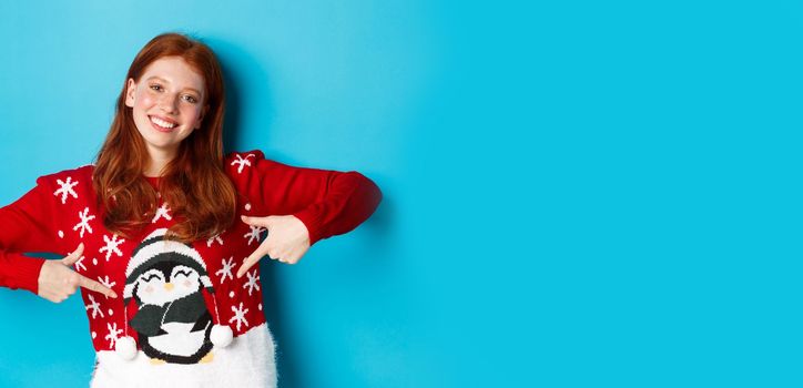 Winter holidays and Christmas Eve concept. Pretty redhead girl pointing fingers at cute xmas sweater with penguin, standing over blue background.