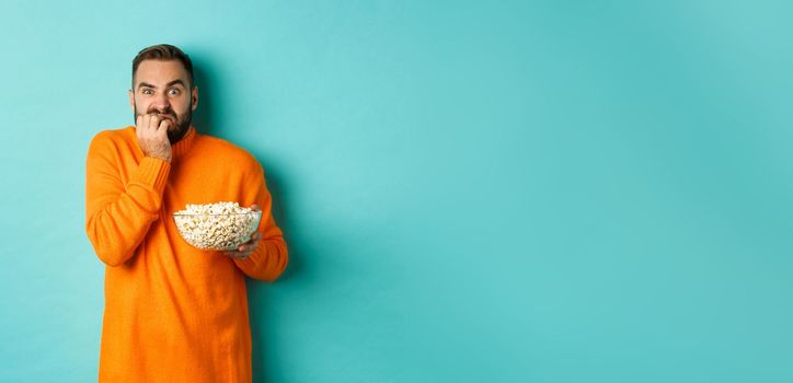 Image of scared young man watching horror movie, biting fist and looking terrified, holding bowl of popcorn, standing over turquoise background.