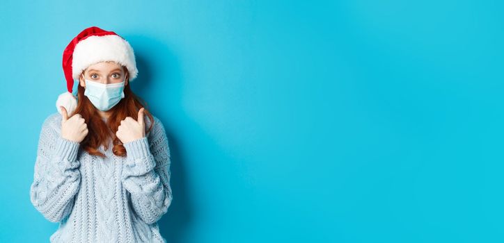 Christmas, quarantine and covid-19 concept. Cute teen redhead girl in santa hat and sweater, wearing face mask from coronavirus, showing thumbs up, standing over blue background.