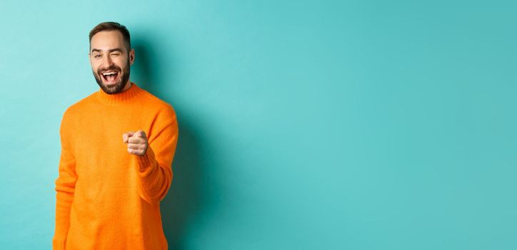 Cheeky attractive man congrats you, praising good job, winking and pointing at camera, standing over light blue background.