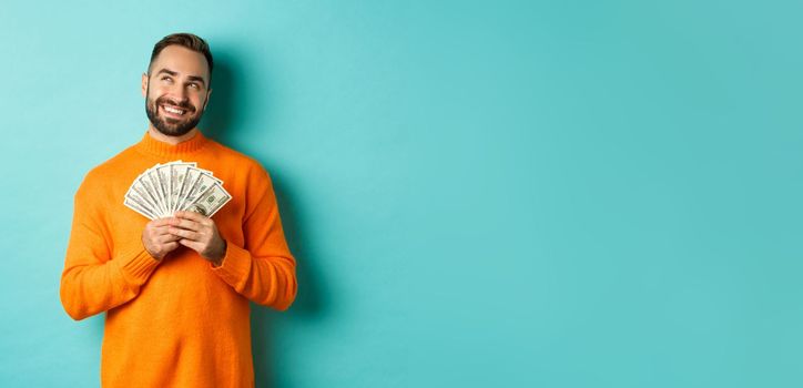 Man thinking about shopping, holding money and smiling satisfied, looking at upper left corner, standing over light blue background.