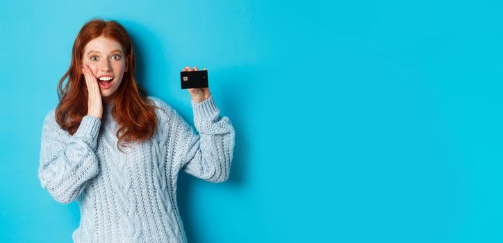 Cute redhead girl in sweater showing credit card, smiling at camera, standing over blue background.