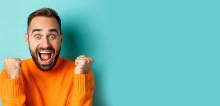 Image of handsome relieved man feeling satisfaction, rejoicing of winning or achievement, making fist pump and saying yes, standing over turquoise background.