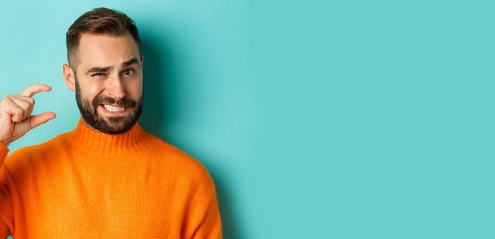 Close-up of awkward man showing small or little with fingers, standing displeased over light blue background.