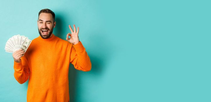 Shopping. Excited guy holding money, showing ok sign and winking, standing over light blue background. Copy space