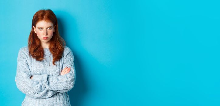 Image of angry redhead girl feeling offended, cross arms on chest and sulking, staring at camera mad, standing against blue background.