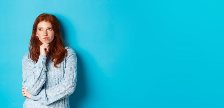 Pretty teenage girl with red hair thinking, looking upper left corner thoughtful, pondering something, standing over blue background.
