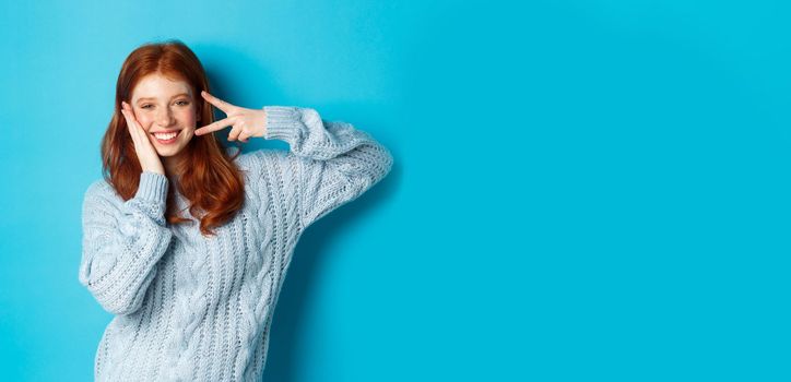 Cheerful redhead female model sending good vibes, smiling and showing peace sign, standing over blue background.