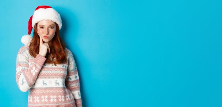 Winter and Christmas Eve concept. Thoughtful redhead teen girl looking at camera, making choice or thinking, wearing santa hat, standing over blue background.