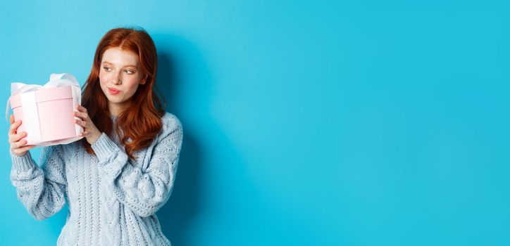 Curious teen girl with red hair, shaking gift box and wonder what inside, standing over blue background.