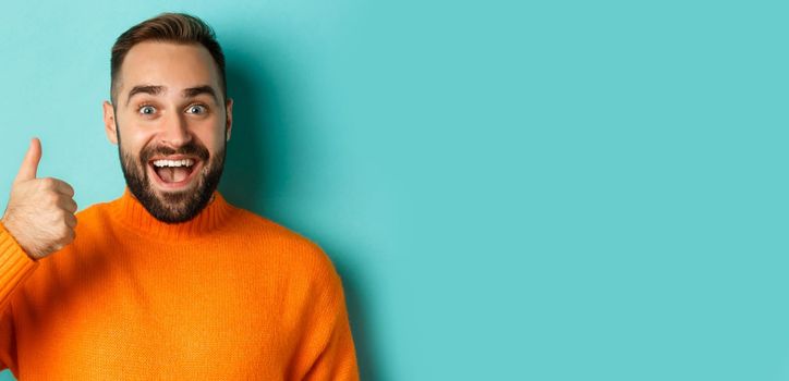 Close-up of cheerful male model showing thumb-up, smiling pleased, standing over light blue background.